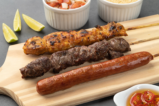 Meat, chicken and pork sausage skewers over wooden board with farofa and vinaigrette.