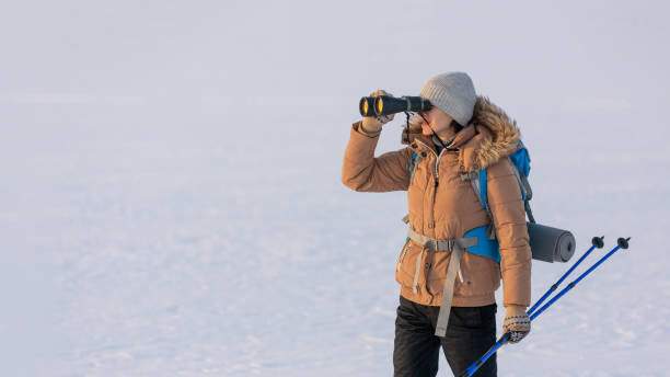 istock Woman in down jacket with a backpack and ski poles in hand looks through binoculars standing on a snowy plain in winter, selected focus. 1386249343