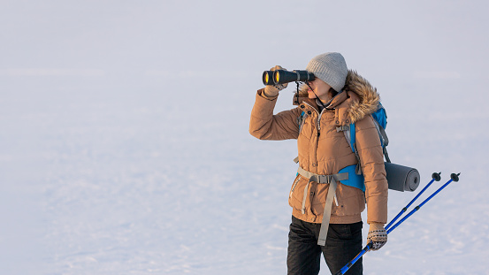 Woman in down jacket with a backpack and ski poles in hand looks through binoculars standing on a snowy plain in winter, selected focus. Hiker, pioneer, adventure, exploration, searches, scientist, extreme travel concept.