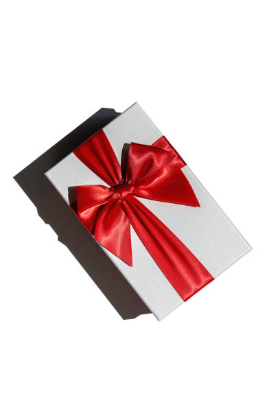 Square closed gift box made of white paper, tied up with wide shiny red satin ribbon and bow isolated with hard shadow on white background. View from directly above. Birthday or Christmas present.