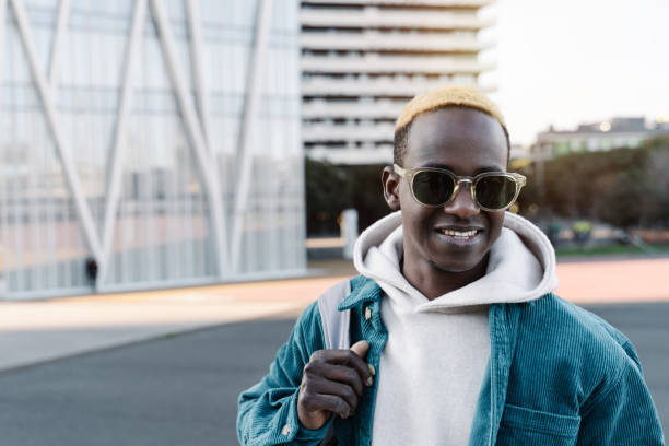 Smiling young African man with blond hair and sunglasses walking with bag in modern city Smiling young African man with blond hair and sunglasses walking with bag in modern city black men with blonde hair stock pictures, royalty-free photos & images