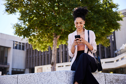 Young businesswoman texting on her phone and smiling while sitting on a bench in front of an office building in summer