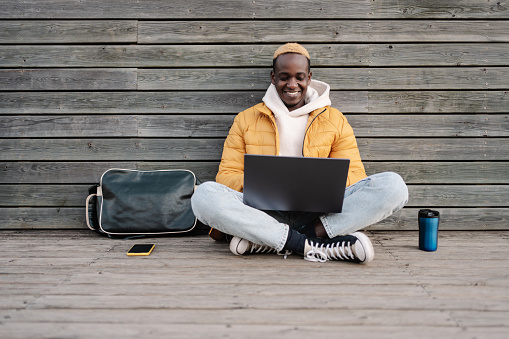 African American male university student sitting outdoors working on laptop smiling wearing yellow hoodie