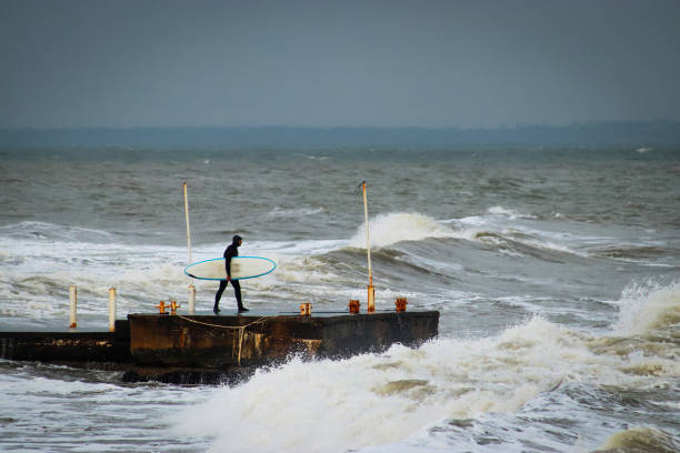 Surfer in a wetsuit with a surfboard on a city pier amidst powerful waves. Concept: confrontation between the elements and humans, extreme sports and hobbies, nature versus humans, surfing as a lifestyle surfista en traje de neopreno con una tabla de surf en un muelle de la ciudad en medio de poderosas olas. concepto: confrontación entre los os y los humanos, deportes extremos y pasatiempos, naturaleza versus humanos, el surf como estilo de vida