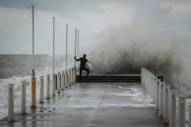istock Alone surfer in wetsuits with surfboard on a city pier amid powerful waves. Concept: confrontation between the elements and man, extreme sports and hobbies, nature vs man. Back view 1386271674