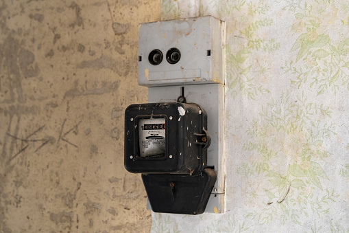 Old electric meter on a wall in an abandoned building. Electrical equipment to measure power consumption for accounting. The vintage wallpaper with dirt spots is in very bad condition.
