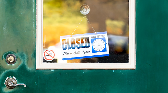 Closeup closed sign hanging and some white paper notice stick on a glass window of weathered green front door. Antique shop temporary shut during crisis. Break time hour for vintage retail business.