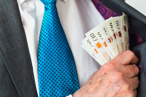 Close up color image depicting a senior businessman putting a wad of cash - paper currency - in the inside pocket of his business suit.