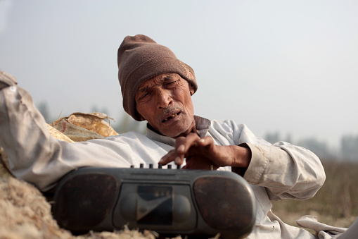 Old farmer of Indian ethnicity sitting portrait outdoor in nature with tape recorder.