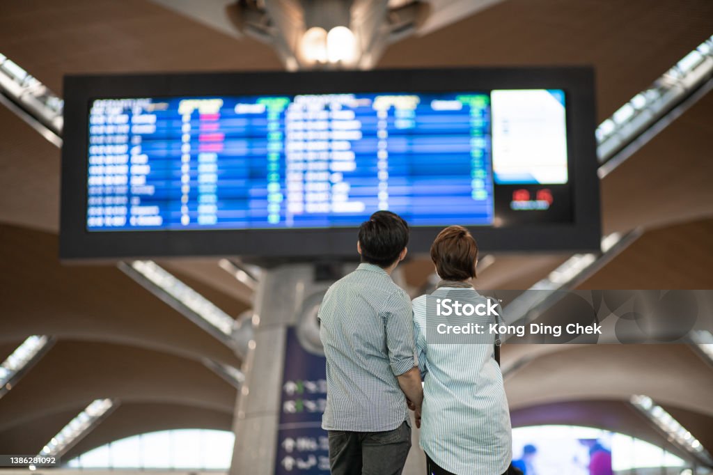 Rear view. Asian Chinese couple checking boarding time in airport.  Waiting in front arrival departure board. Flying Stock Photo