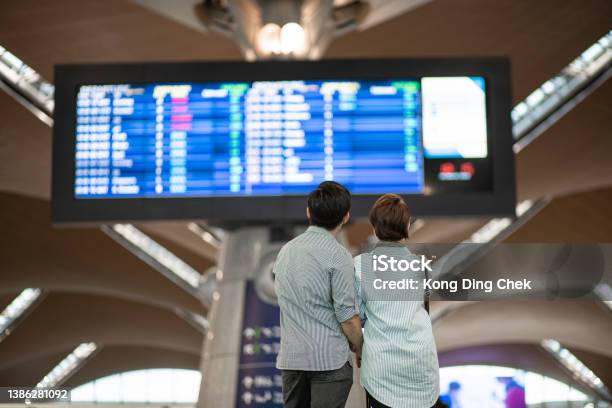 Rear View Asian Chinese Couple Checking Boarding Time In Airport Waiting In Front Arrival Departure Board Stock Photo - Download Image Now