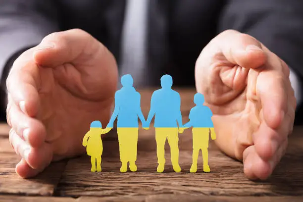 Close-up Of A Businessperson's Hand Protecting Family Figures On Wooden Desk