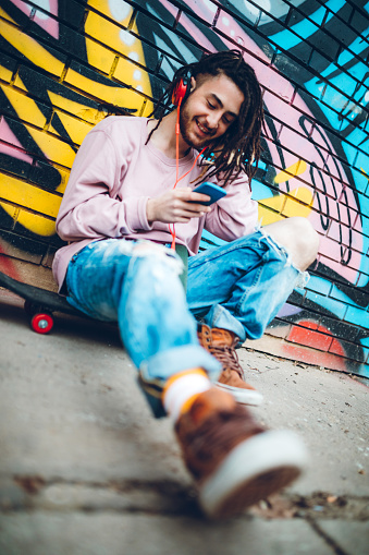 Young caucasian graffiti artist listens to music and using phone while sitting on a skateboard.