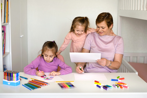 Grandma helps granddaughter draw a drawing with pencils and felt-tip pens. Kids have fun with Grandmother. Grandparents teach grandchildren to draw correctly and beautifully. Home preschool education