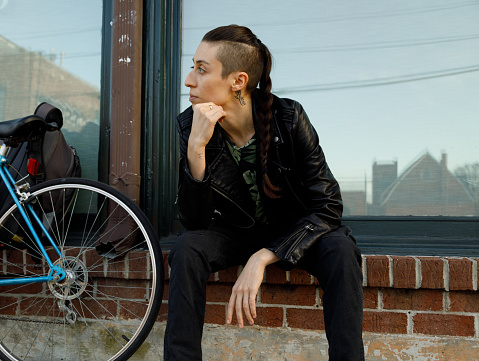 A transgender person sits against a building in Atlanta, Georgia.  They are wearing a black leather jacket and black pants with white sneakers.  They have dark hair that is shaved on the sides and worn in a ponytail.  Their bike is leaning against the wall next to them.  They are looking off camera and resting their face in their hand.