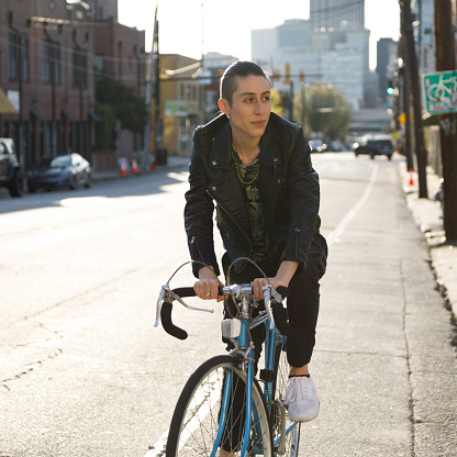A transgender person rides a bike on the street in Atlanta, Georgia.  They are wearing a black leather jacket, black pants, and white sneakers.  They are on a blue road bike.  They have dark hair that is shaved on the sides and worn in a ponytail.  It is last afternoon and the sun is behind the subject.