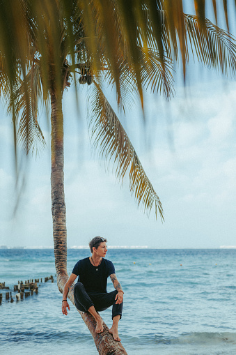 An urban young man is sitting on a palm tree on a beautiful tropical beach