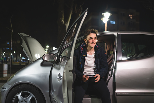 One young adult caucasian man sitting his vehicle with open door and hood and broken failed engine holding a phone calling towing service for help on the road Roadside assistance concept in dark night