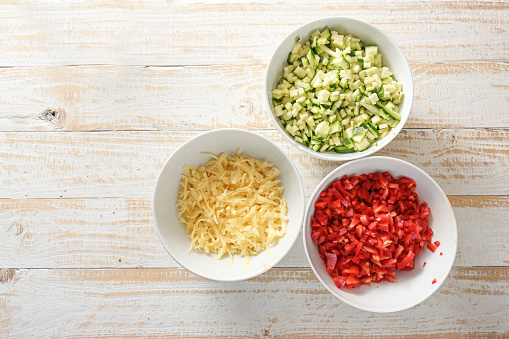 Diced vegetables, zucchini and red bell pepper, and grated cheese in white bowls on a rustic wooden table, copy space, high angle view from above, selected focus