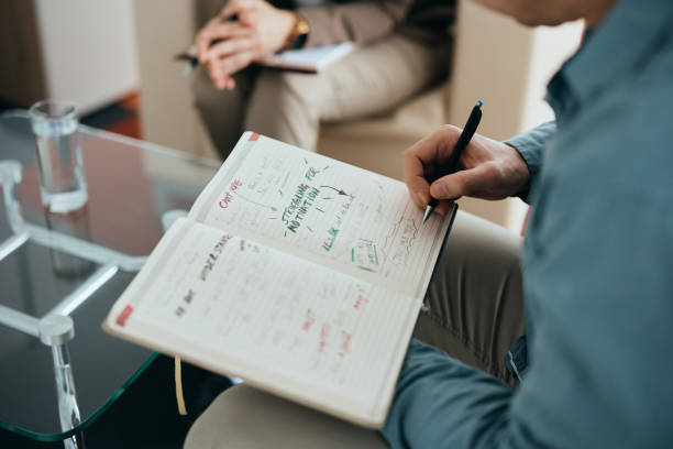 Close-up of man brainstorming during therapy with his psychiatrist. Close-up of male patient writing in notebook while brainstorming during counseling with mental health professional. personality test stock pictures, royalty-free photos & images