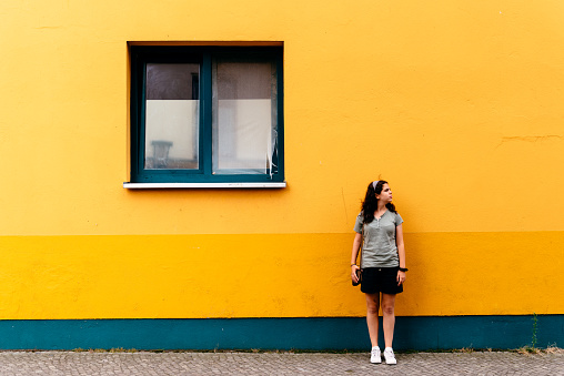 Young woman leaning against a bright wellow wall. She is confident and happy