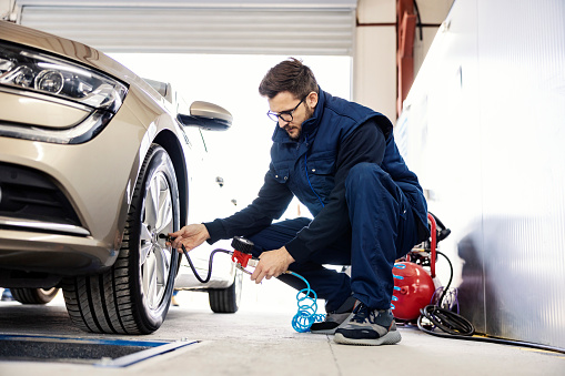 A mechanic crouching next to a car and inflates the tire at workshop.