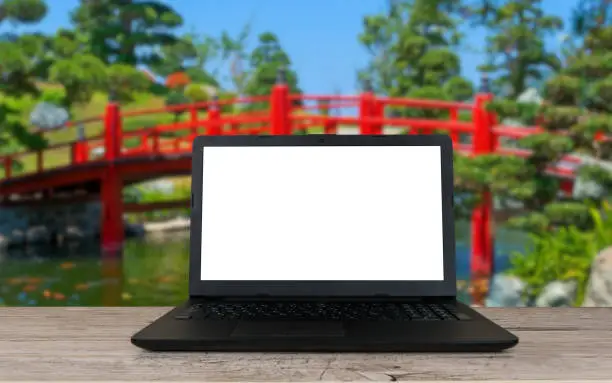 open laptop with blank screen on wooden table and red bridge in japanese garden blurred background, remote or distant work place, travel or vacation working concept