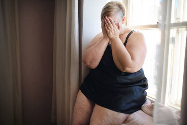 подавленная одинокая толстая женщина сидит у окна дома и закрывает лицо. - mature adult sadness overweight women стоковые фото и изображения