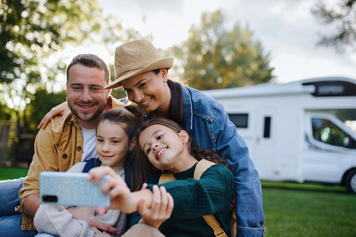 A happy young family with two children ltaking selfie with caravan at background outdoors.