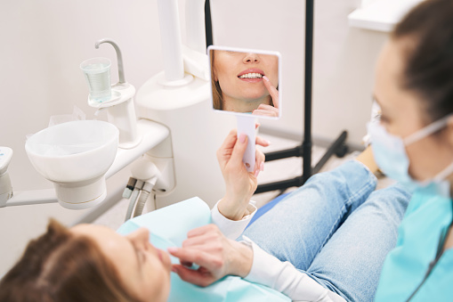 Close up of female person lying in dental chair and looking at teeth reflection while discussing dental treatment with dentist