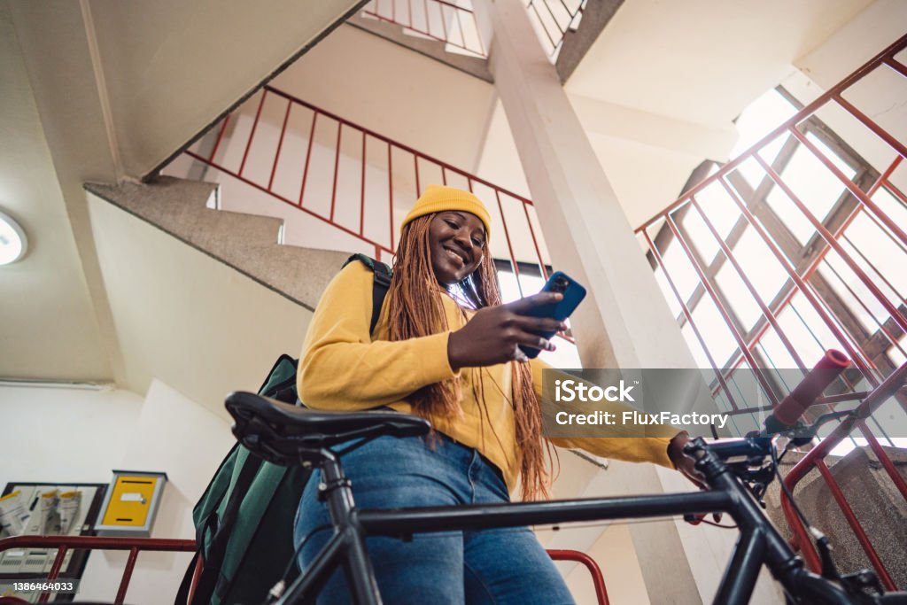 female bicycle delivery person Black ethnicity using mobile phone, in the building Low angle view of female bicycle delivery person Black ethnicity using mobile phone, in the building Bicycle Stock Photo