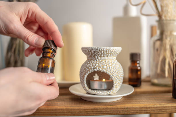 La main de la femme tient une bouteille d’huile essentielle et ouvre le couvercle. Il y a une lampe aromatique, des bougies et des cosmétiques sur la table. Concept d’aromathérapie, atmosphère relaxante et rituels - Photo