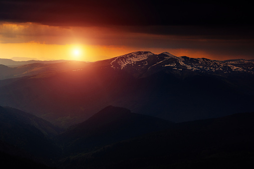 Scenic landscape in the mountains at sunset. View of dramatic sky over snow-capped mountain range covered by snow residual snow in early spring  in background. Dusk scene.