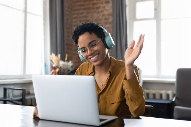 Cheerful happy African American hipster girl in headphones and glasses Cheerful happy African American hipster girl in headphones, glasses waving hand hello at webcam of laptop, smiling, speaking on video conference talk, using computer for remote virtual communication hello stock pictures, royalty-free photos & images