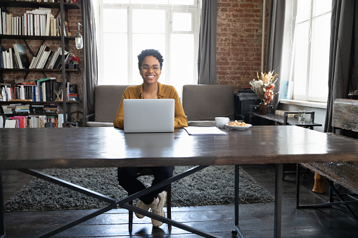 Happy African American freelance, worker, entrepreneur, student using laptop at wide wooden table workplace, looking at camera, smiling, laughing. Business woman in home office full length portrait