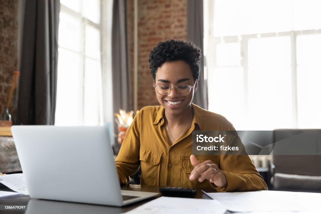 Happy young Afro American entrepreneur woman in glasses counting profit Happy young Afro American entrepreneur woman in glasses counting profit, on calculator at laptop computer, analyzing benefits, enjoying financial success, job high result, smiling Finance Stock Photo