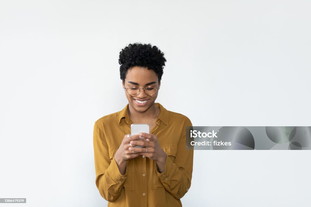 Happy Black woman in casual glasses browsing internet Happy millennial Black woman in casual glasses browsing internet on mobile phone, reading, typing text message, chatting online, smiling, laughing, using banking app on smartphone. Isolated portrait Using Phone Stock Photo