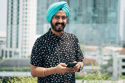Cheerful smiling business man with turban typing text message on his phone and looking at camera while standing in the city.