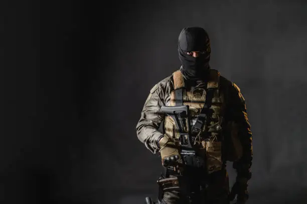 Studio portrait of a male soldier with a rifle wearing balaclava, he is standing against black background.