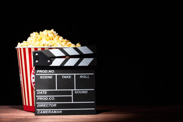 Cinema concept. Popcorn in a box and movie clapper on wooden table under beam of light against black background Cinema concept. Popcorn in a box and movie clapper on wooden table under beam of light against black background. pop art photos stock pictures, royalty-free photos & images