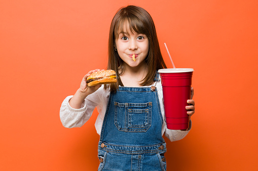 Adorable Caucasian girl enjoying a burger combo with a large soda and some fries in her mouth making a funny face