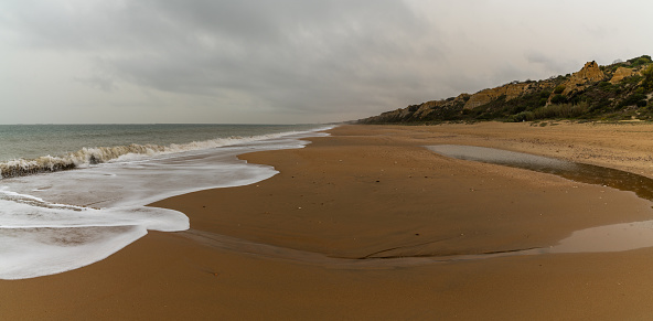 A panorama view of a beautiful long and empty beach with shore break and high sand dunes behind