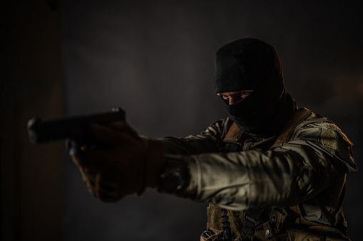 Studio portrait of a male soldier wearing balaclava and pointing with a handgun, he is standing against black background.
