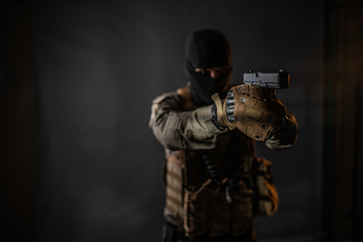 Studio portrait of a male soldier wearing balaclava and pointing with a handgun, he is standing against black background.