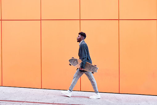 side view of a young man with blazer and skateboard walking down the street against an orange background.