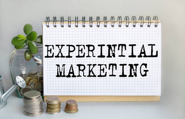 istock EXPERINTIAL MARKETING is written in a notebook that sits on the table along with a stack of coins, a glass jar with coins and a sprout, and a small toy watering can. Money saving concept. 1386524572