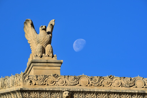An eagle sculpture on the top of a federal building spreads its wings next to a gibbous moon in Washington, D. C. on a clear blue-sky day.