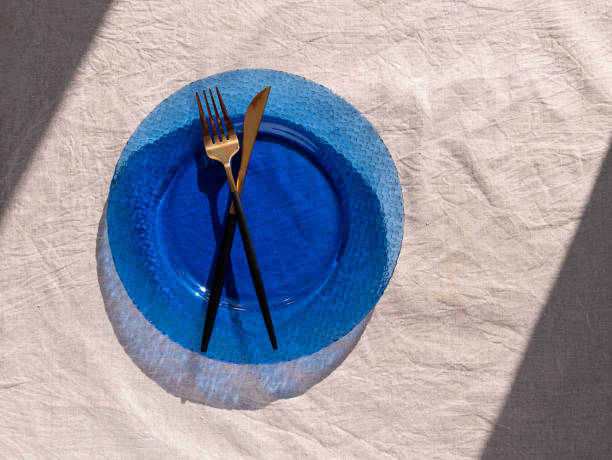 istock Table setting empty blue glass plate with fork knife on linen cloth top view daylight harsh shadows. Festive dish place. 1386548630