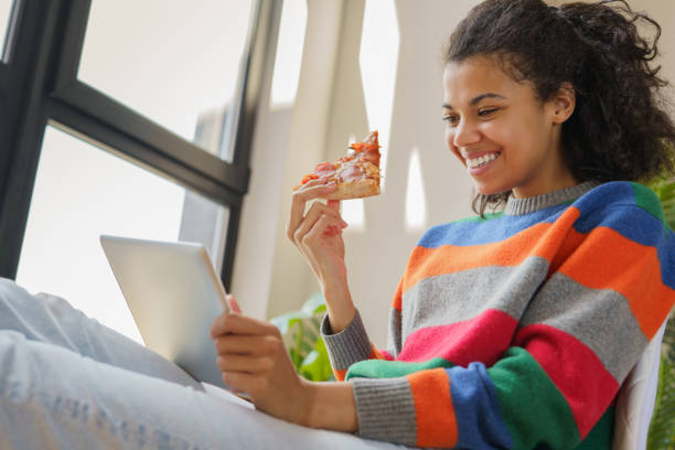 Smiling African American woman using digital tablet watching online movie, eating pizza relaxing at home stock photo