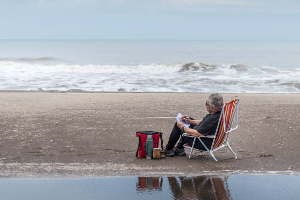 uomo adulto con capelli grigi e occhiali seduto su una sedia a sdraio che disegna. dietro la spiaggia e le onde del mare. - 4598 foto e immagini stock